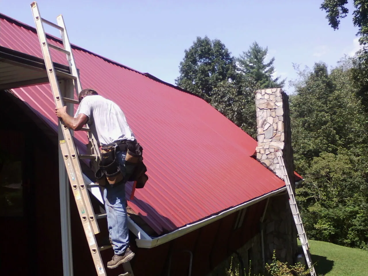 Roofer on a ladder beside a red metal roof and stone chimney — Soffit & Fascia Repair by Roofing Brothers in Stowell